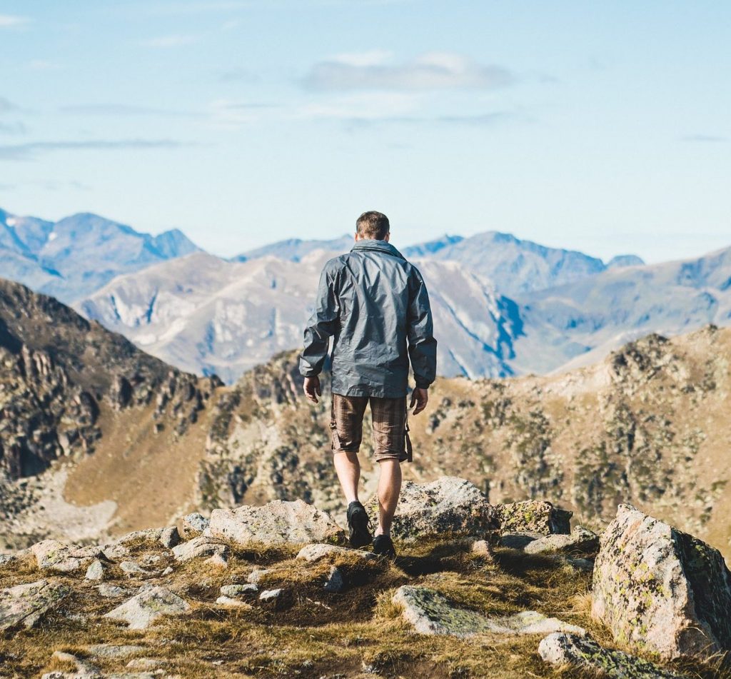 Ein Mann, von Hinten zu sehen, steht auf einem Fels und blickt auf das Gebirge unter ihm.