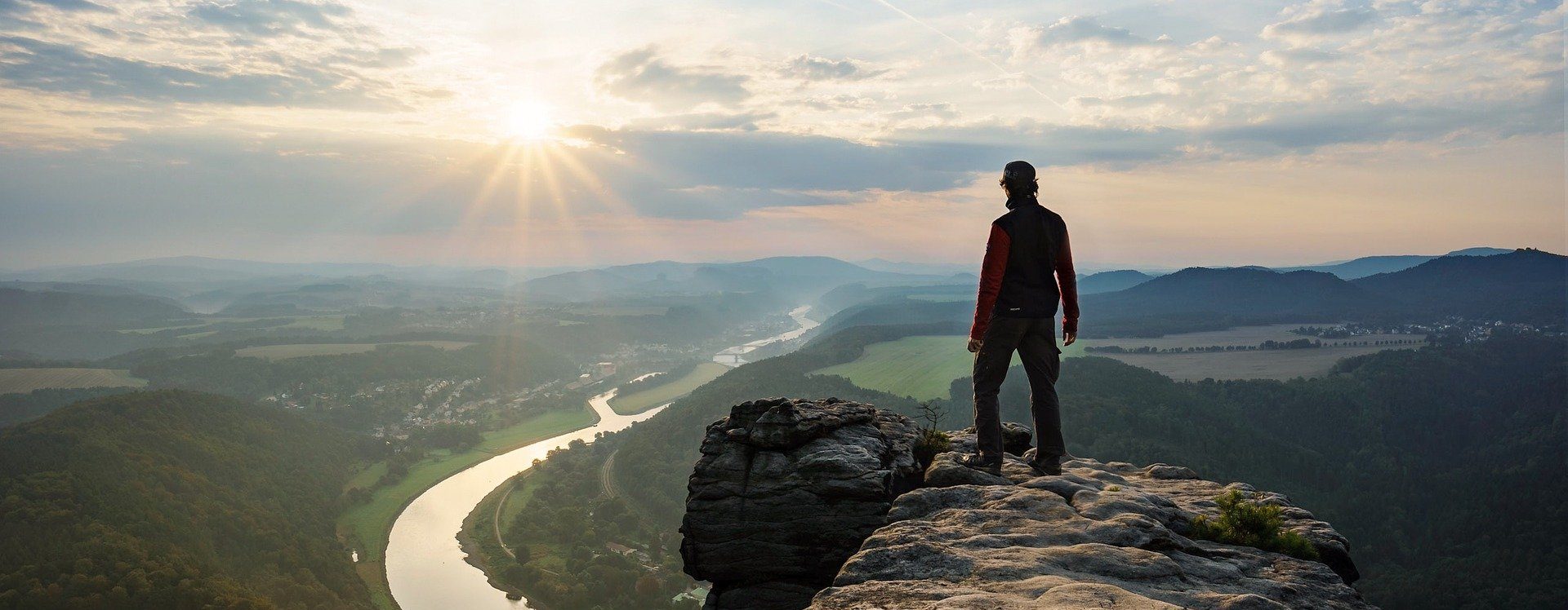 La silueta de un montañero en la puesta de sol, de pie en el borde de un acantilado y mirando hacia el valle con un río.