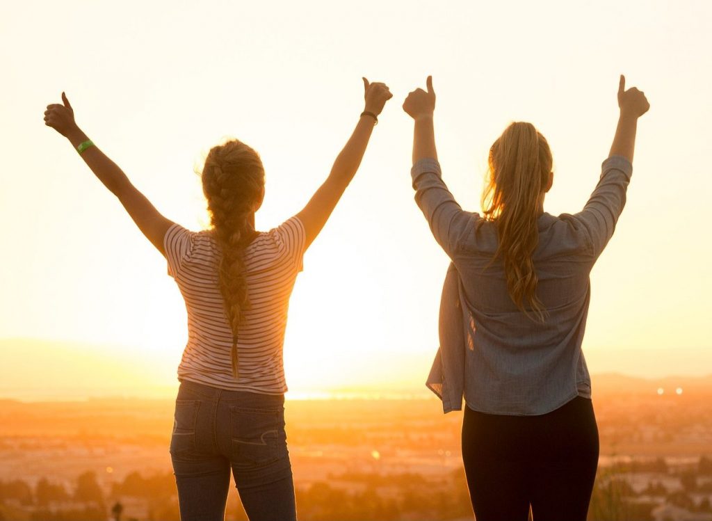 Zwei Frauen stehen mit hochgereckten Daumen auf einem Aussichtspunkt und schauen in den Sonnenuntergang.