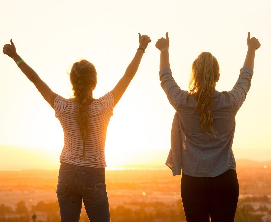 Zwei Frauen stehen mit hochgereckten Daumen auf einem Aussichtspunkt und schauen in den Sonnenuntergang.