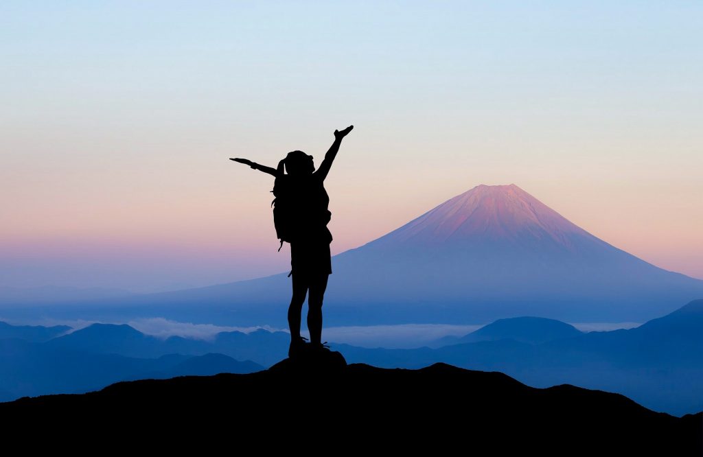 Eine reisende Frau steht auf einem Berg und streckt die Arme in den Himmel bei Sonnenaufgang mit einem Vulkan im Hintergrund