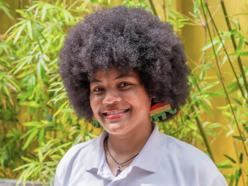 Hotelmitarbeiter*in trägt ein weißes Poloshirt, steht vor Pflanzen und blickt direkt in die Kamera.