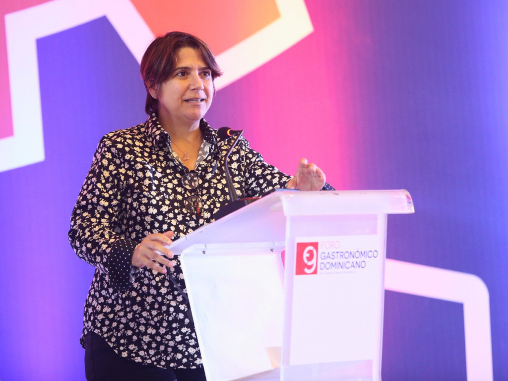 En la imagen, Lisette está de pie detrás de un atril, frente a una pared iluminada con luces naranjas y moradas.