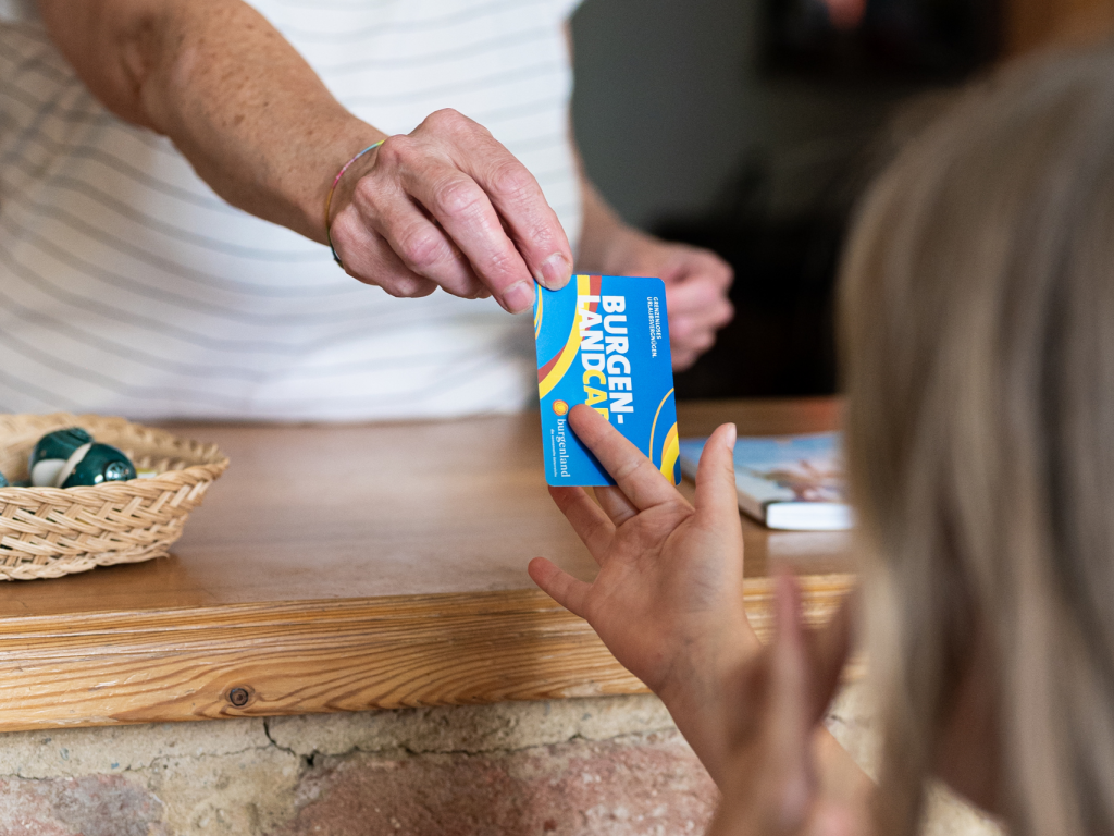 Eine Frau überreicht einer Touristin an einem Stand eine Burgenland Card.